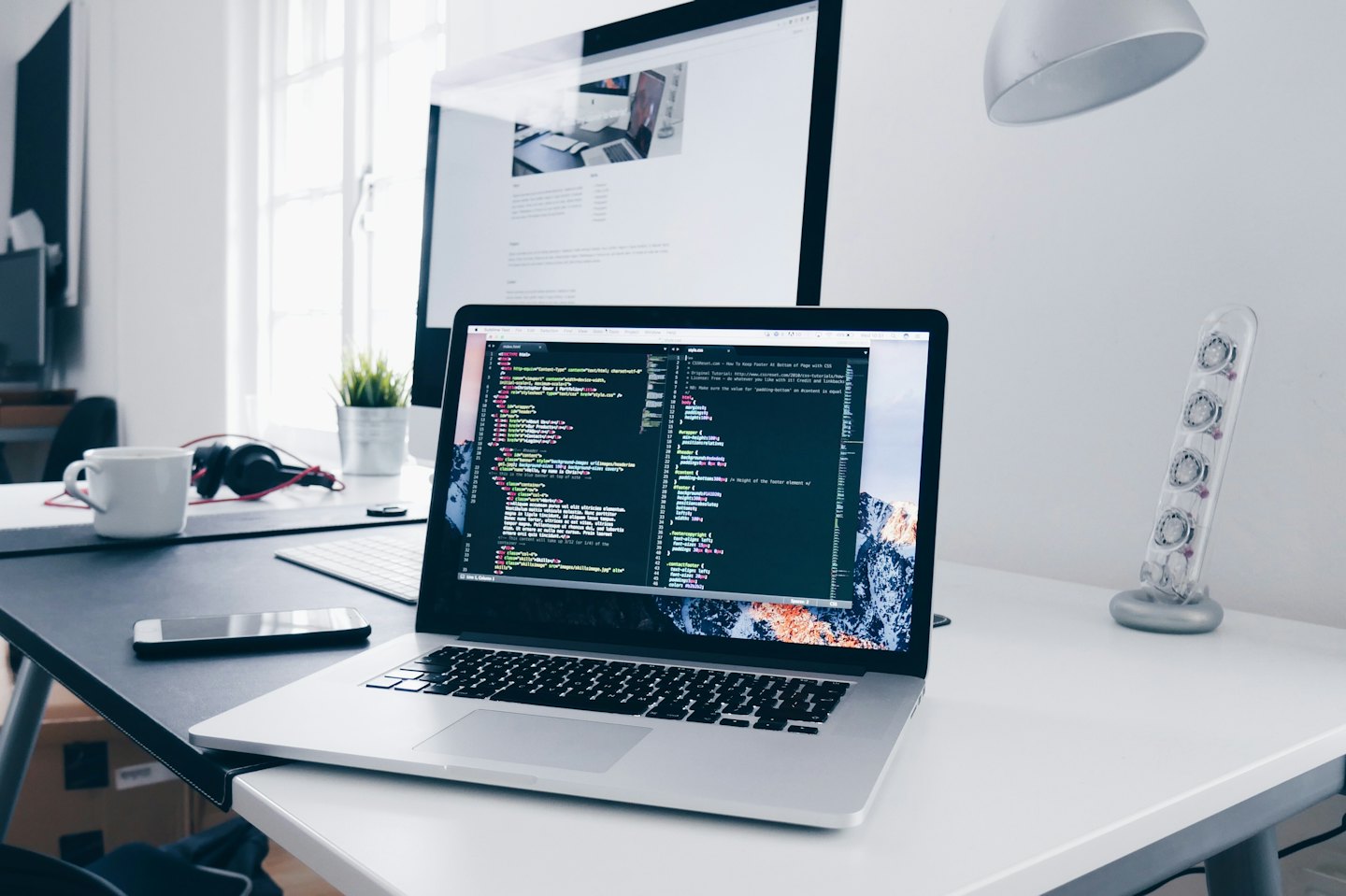 Silver laptop displaying code on a white desk with desktop monitor.